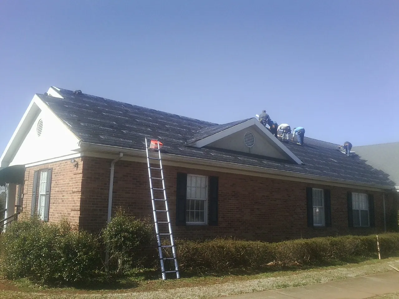 Crew tearing shingles off a brick ranch roof with a ladder set — Emergency Roof Repair work by our crew in Braman