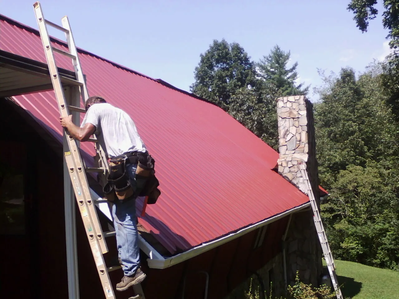 Roofer on a ladder beside a red metal roof and stone chimney — Soffit & Fascia Repair by Roofing Brothers in Braman